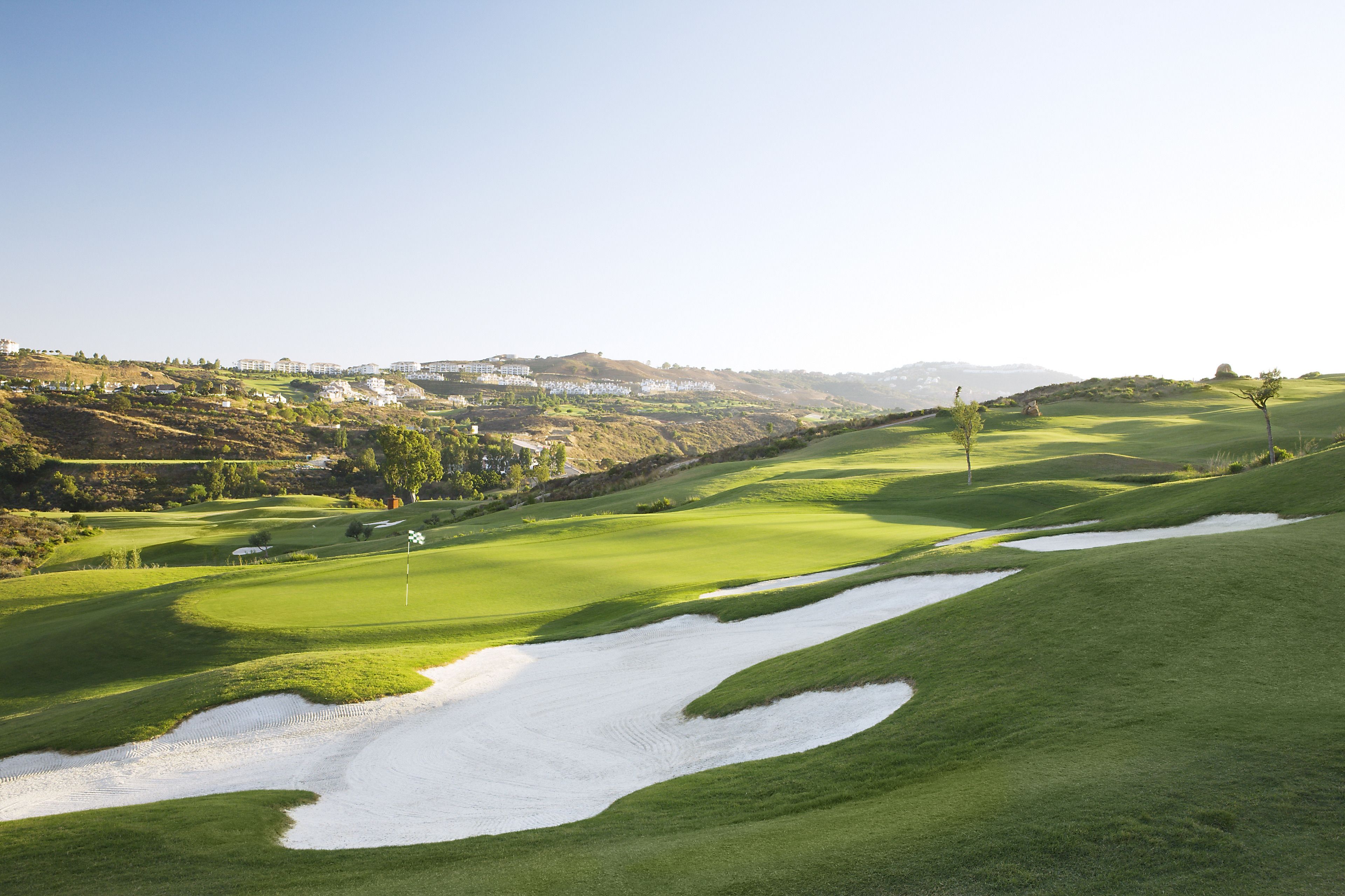 A golf course with bunkers, a flag, and a distant view of buildings.