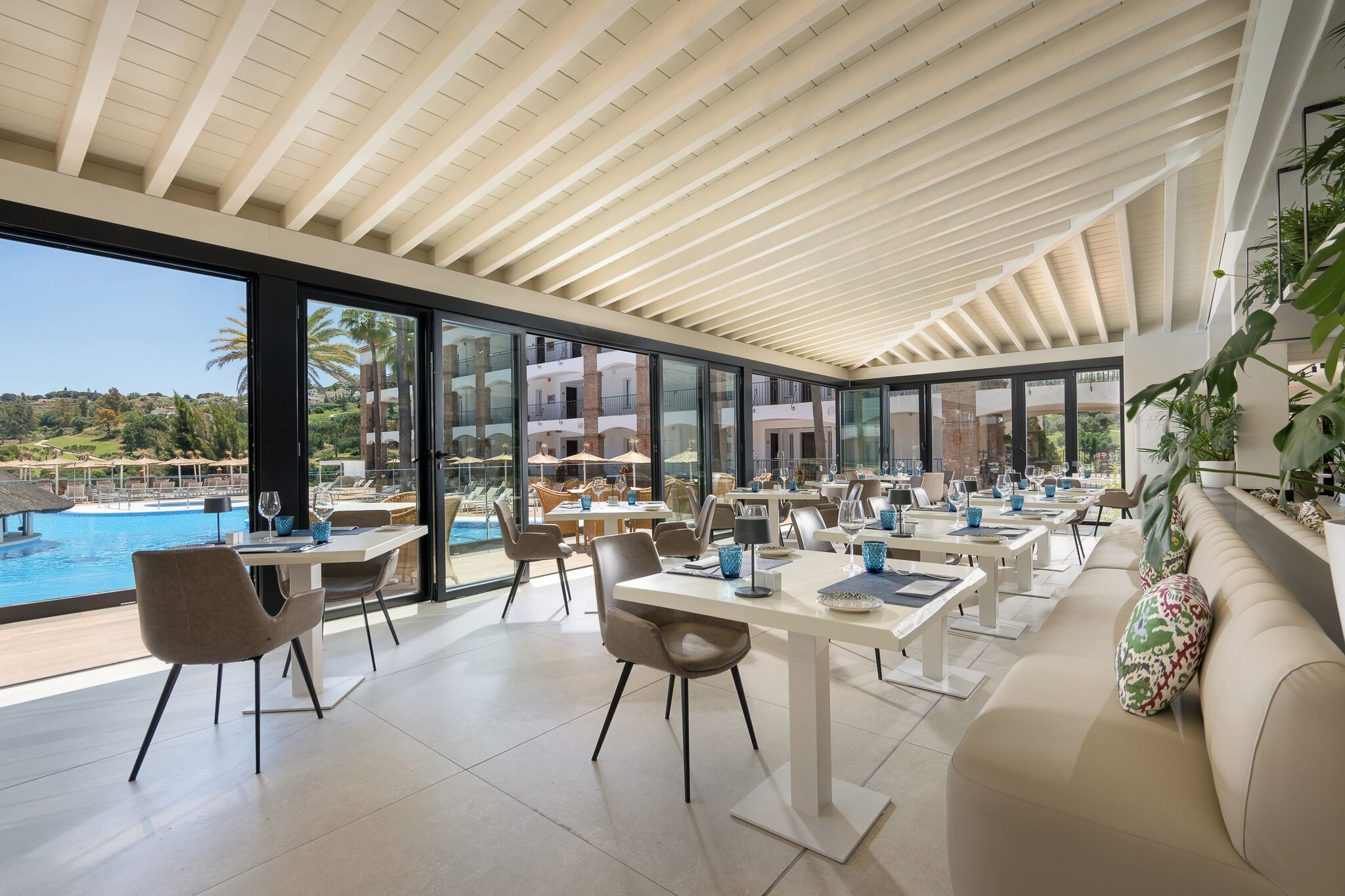 Un espacio interior amplio con mesas y sillas, ventanas grandes y vista a la piscina.