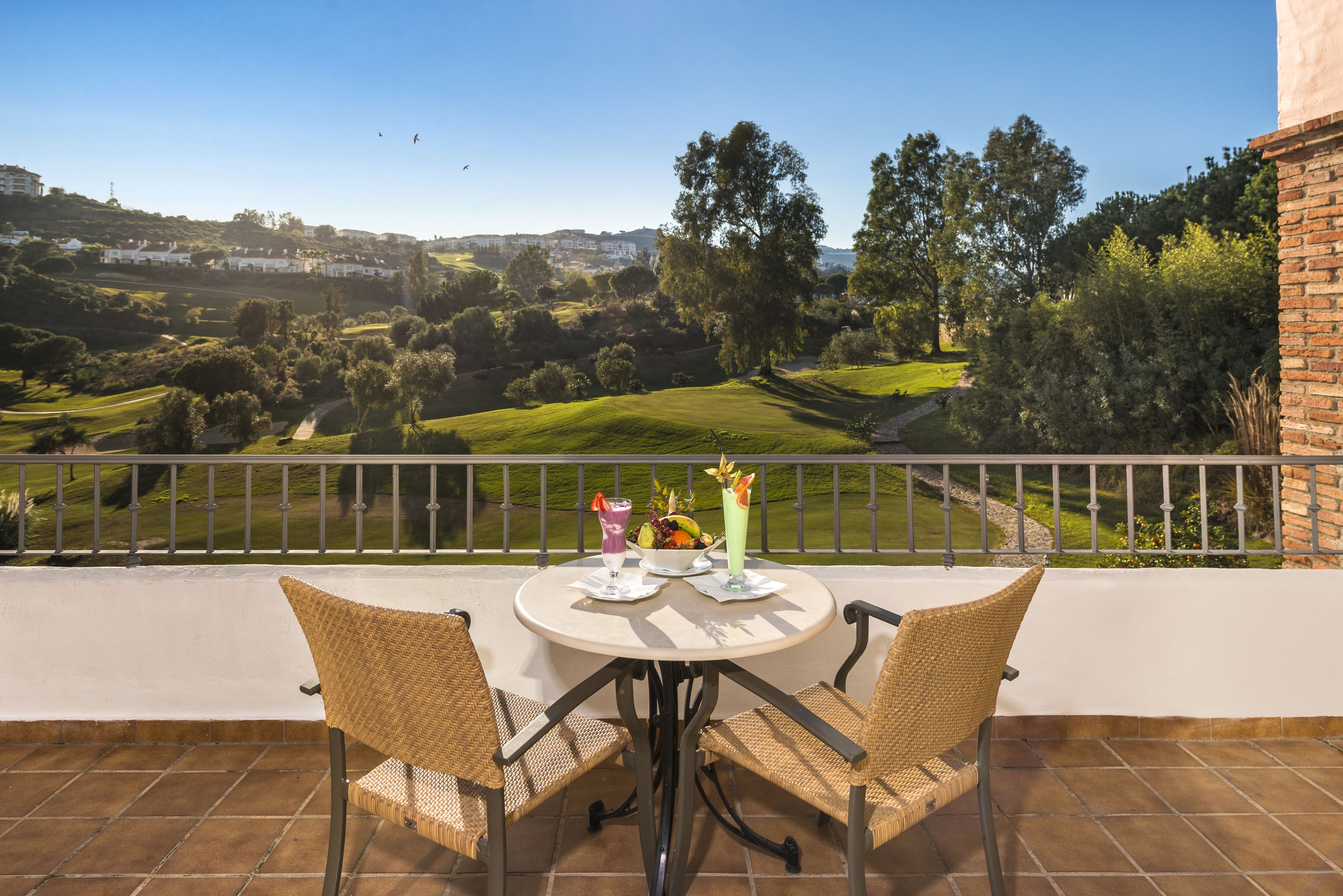 Un balcón con sillas de mimbre y una mesa, con vistas a un campo de golf y edificios residenciales.