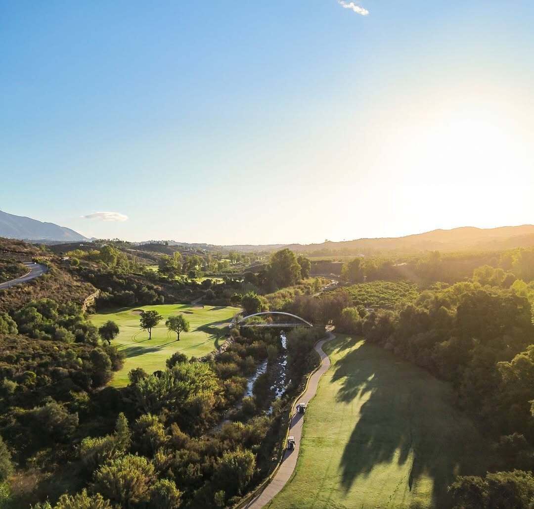Vue aérienne d’un terrain de golf avec un pont, entouré de verdure et de collines.