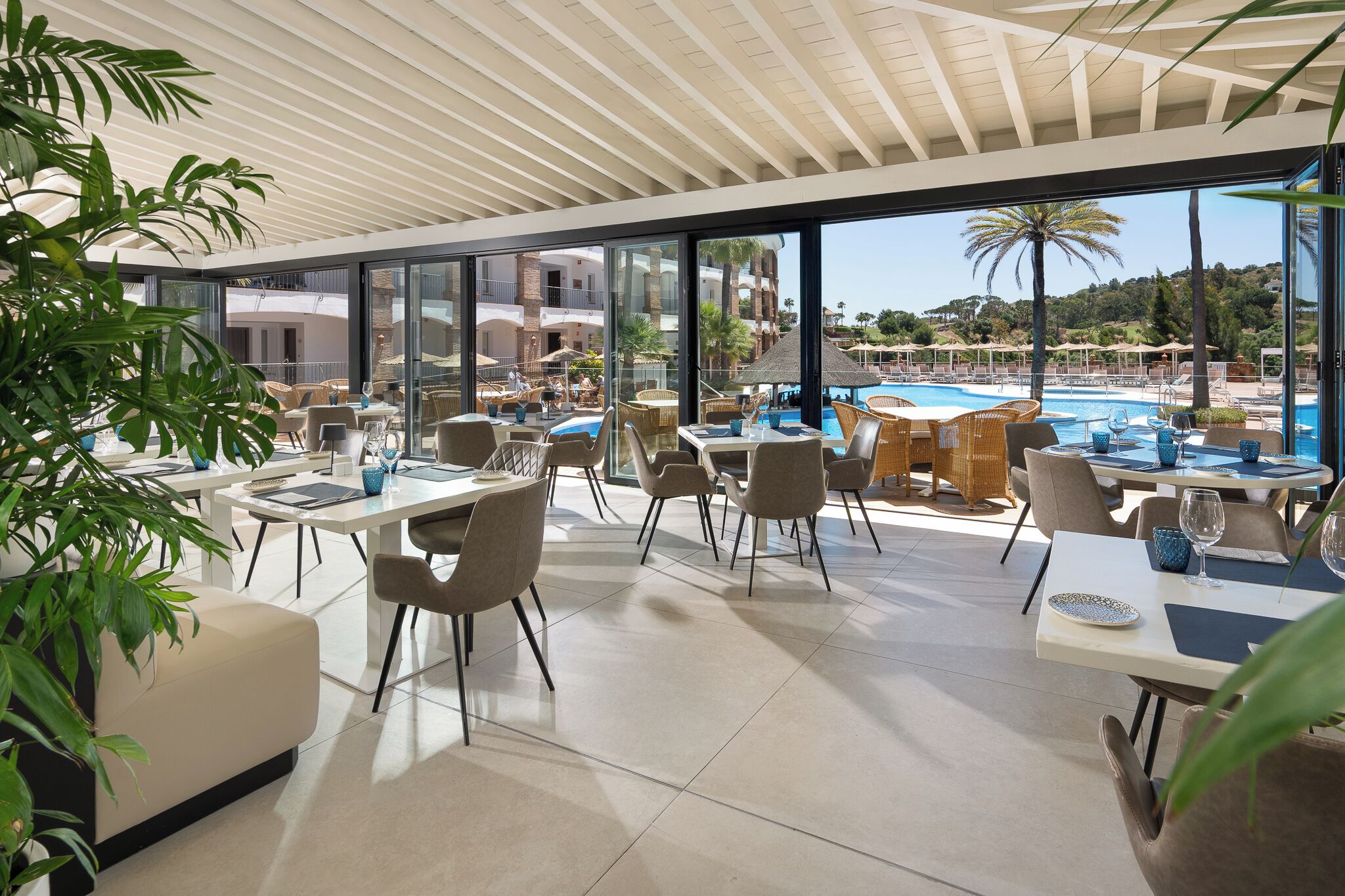 A modern indoor dining area with large windows overlooking a pool and a scenic view of hills.