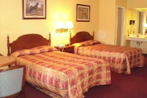 A hotel room with two beds, a bedside table with a lamp, a chair, and a mirror above the sink.