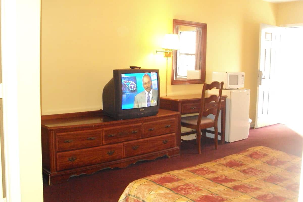 Ein Hotelzimmer mit einem Holzkoffer, einem Fernseher, der eine Nachrichtensendung zeigt, einer Mikrowelle und einem Bett mit einem gemusterten Überwurf.
