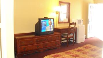 Habitación de hotel con cómoda de madera, televisor mostrando un noticiero, microondas y cama con colcha estampada.