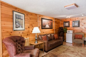 A cozy cabin interior with wooden walls, a leather sofa, a cushioned armchair, a small table with a lamp, and a microwave.