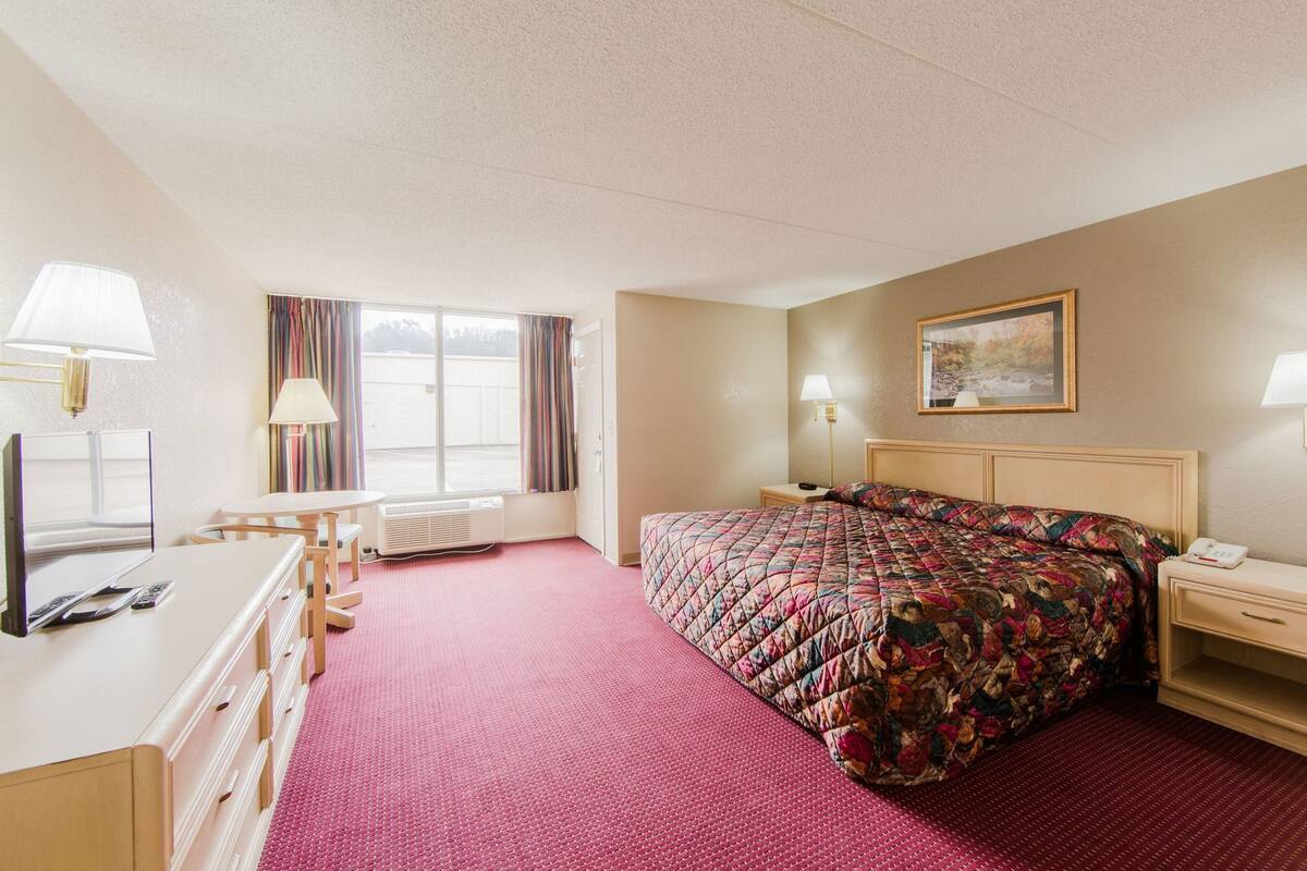 Quarto de hotel com cama, mesa de cabeceira, luminária e janela com cortinas.