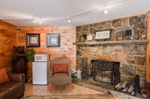 A cozy cabin interior with wooden walls, a stone fireplace, a small refrigerator, and a patterned armchair.