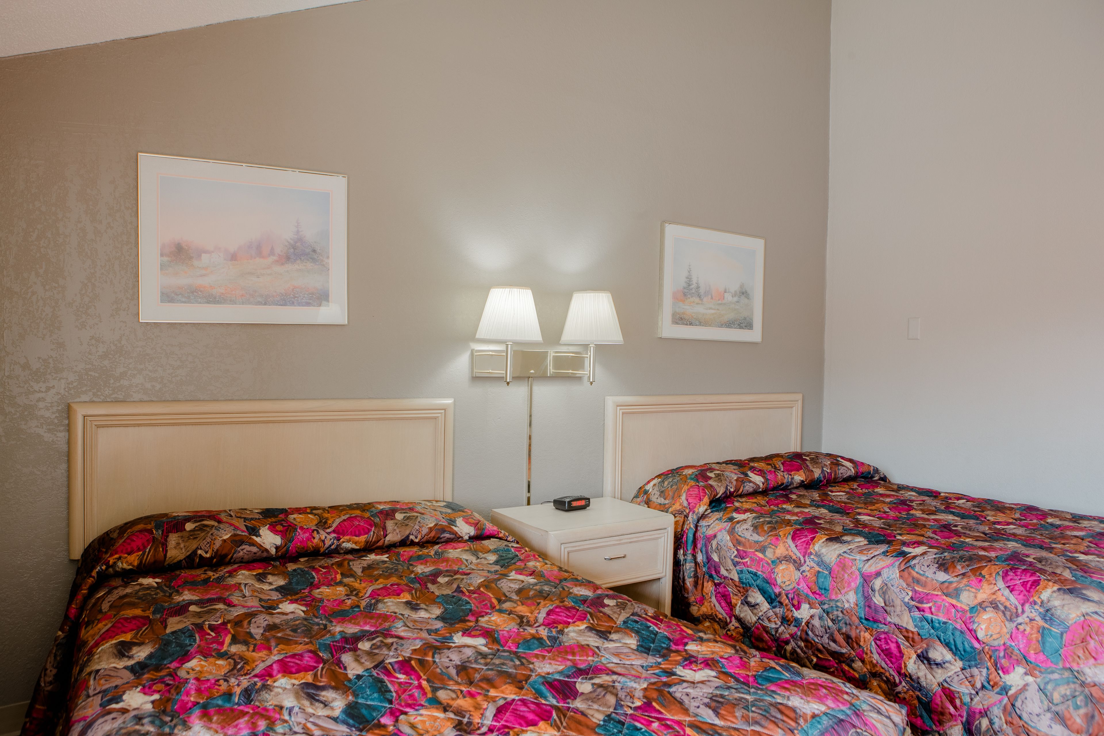 A hotel room with two beds, a nightstand, two wall-mounted lamps, and two framed pictures on the wall.