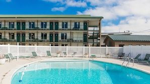 Un espace piscine avec, en arrière-plan, un bâtiment à deux étages, une clôture blanche et plusieurs chaises longues.