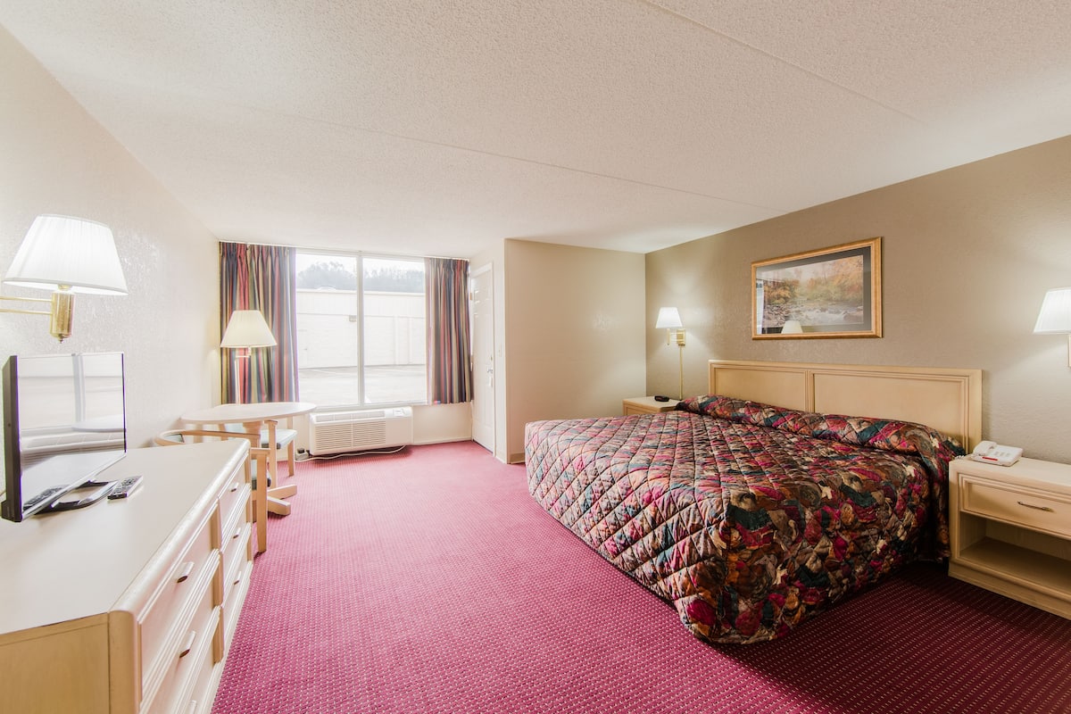 Quarto de hotel com cama, escrivaninha, cadeira, janela com cortinas, quadro na parede e luminária na escrivaninha.