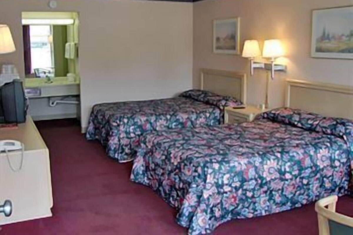 Quarto de hotel com duas camas, uma escrivaninha, uma cadeira, um abajur e um quadro na parede.