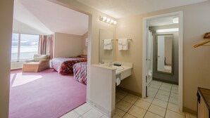 Une chambre avec une grande fenêtre, un lit, une table de chevet et une salle de bain équipée d’un lavabo et d’un miroir.