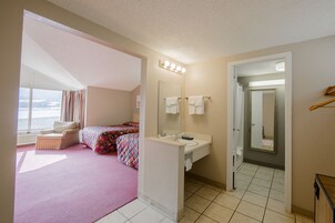 A bedroom with a large window, a bed, a bedside table, and a bathroom with a sink and mirror.