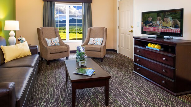 A hotel room with two armchairs, a sofa, a coffee table, a TV on a wooden stand, and a view of a green landscape through a window. - The Resort at Governor's Crossing (Sevierville)