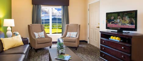 Une chambre d’hôtel avec deux fauteuils, un canapé, une table basse, un téléviseur posé sur un meuble en bois, et une vue sur un paysage verdoyant par la fenêtre.