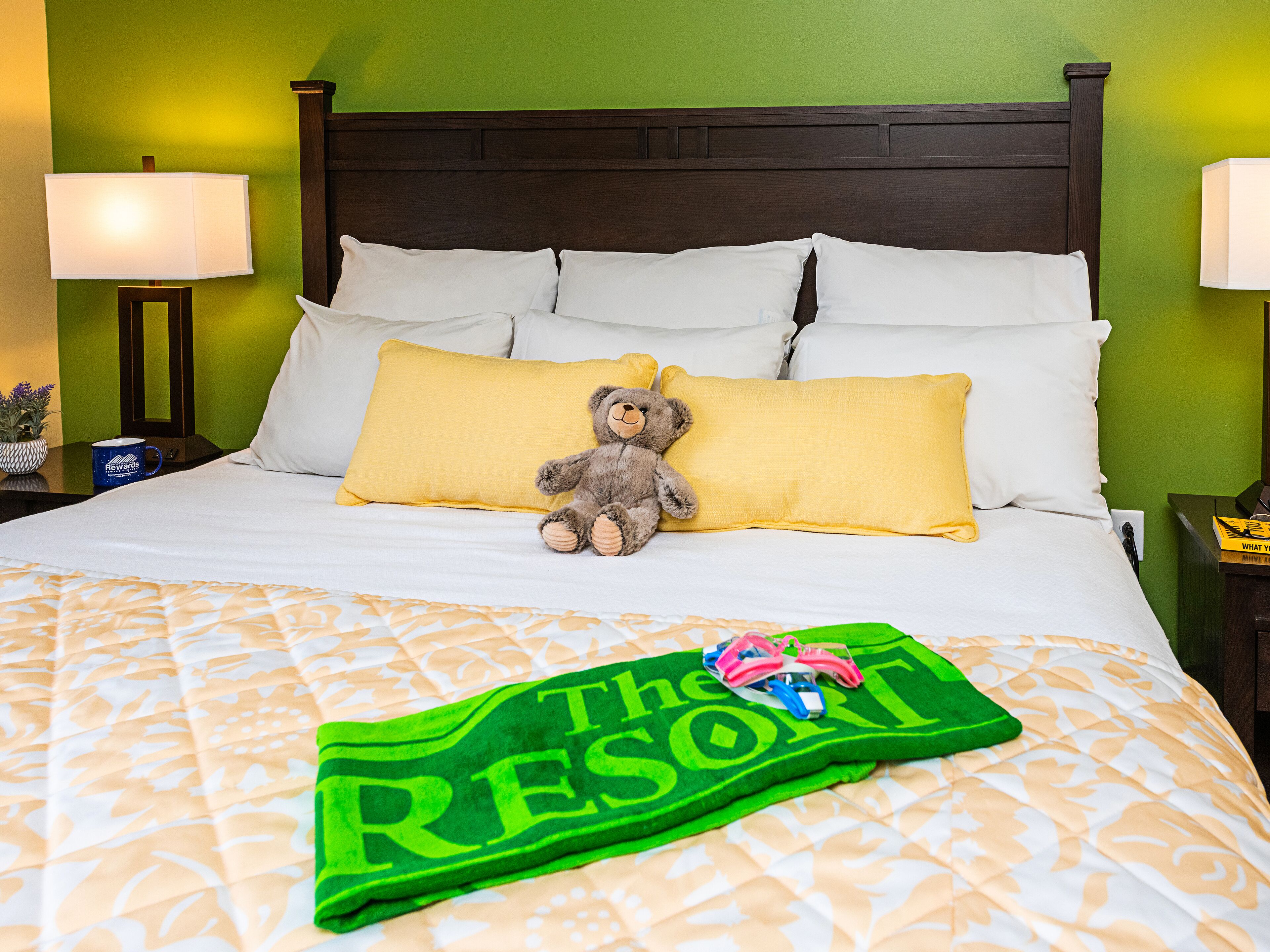 Een net opgemaakt bed met een groene handdoek, een teddybeer, en een groen bordje met daarop 'The Resort'.