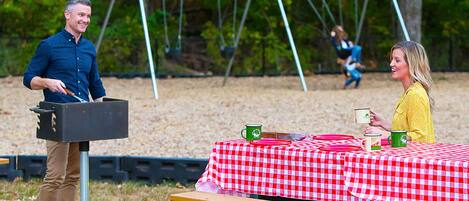 Un montaje para picnic con mantel a cuadros, una parrilla y un conjunto de columpios al fondo.