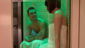Una sauna con dos personas sentadas en bancos, piso de madera y techo con luces.