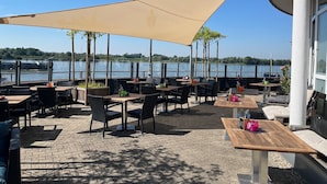 An outdoor patio with tables and chairs, a large canopy, and a view of a lake. - Hotel Rheinpark Rees (Rees)