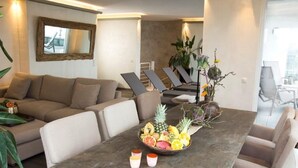 A modern living room with a sofa, dining table with fruit and drinks, and a view of the city through large windows. - Hotel Rheinpark Rees (Rees)
