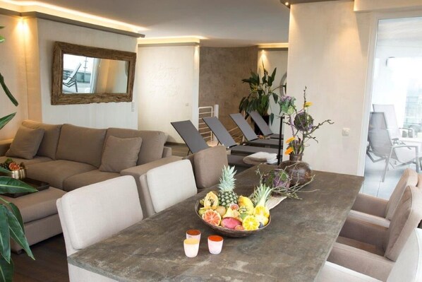 A modern living room with a sofa, dining table with fruit and drinks, and a view of the city through large windows.