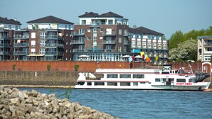 A riverfront hotel with multiple balconies, a white boat docked nearby, and a brick building in the background. - Hotel Rheinpark Rees (Rees)