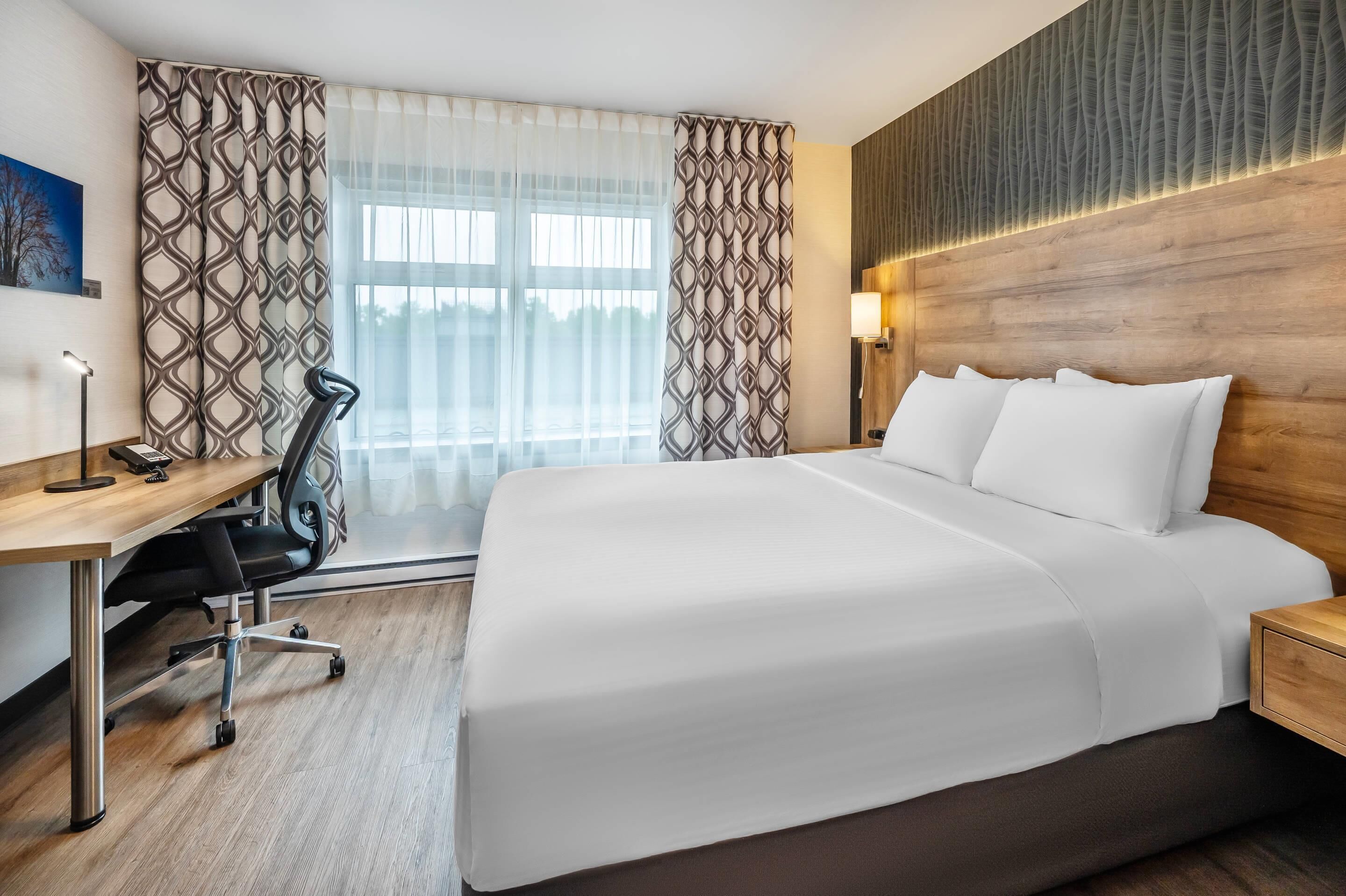 Habitación de hotel con una cama grande, un escritorio con silla, una lámpara y vistas al exterior a través de cortinas traslúcidas.