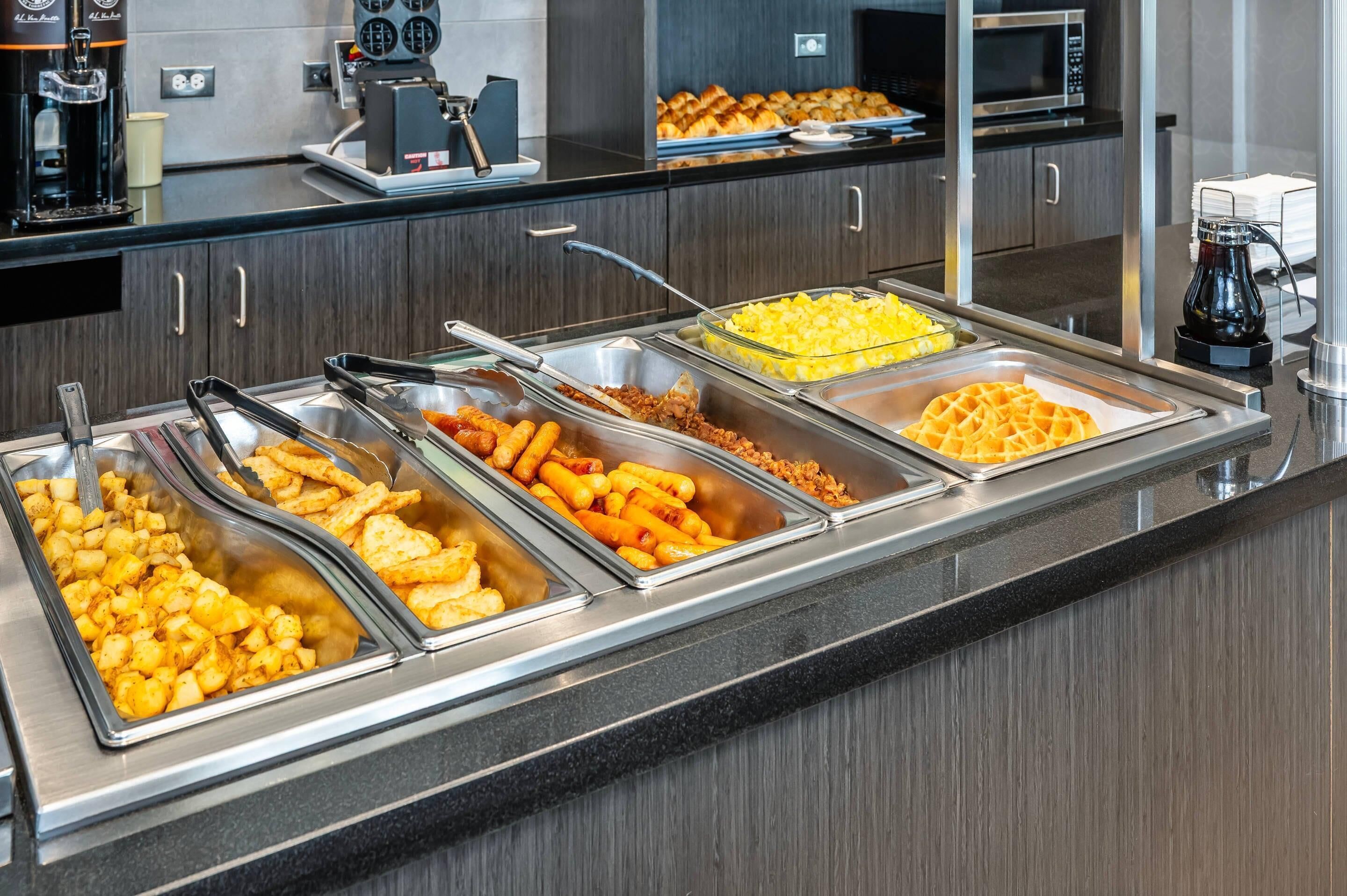 A buffet with various food items including fried potatoes, sausages, waffles, and a fruit salad.