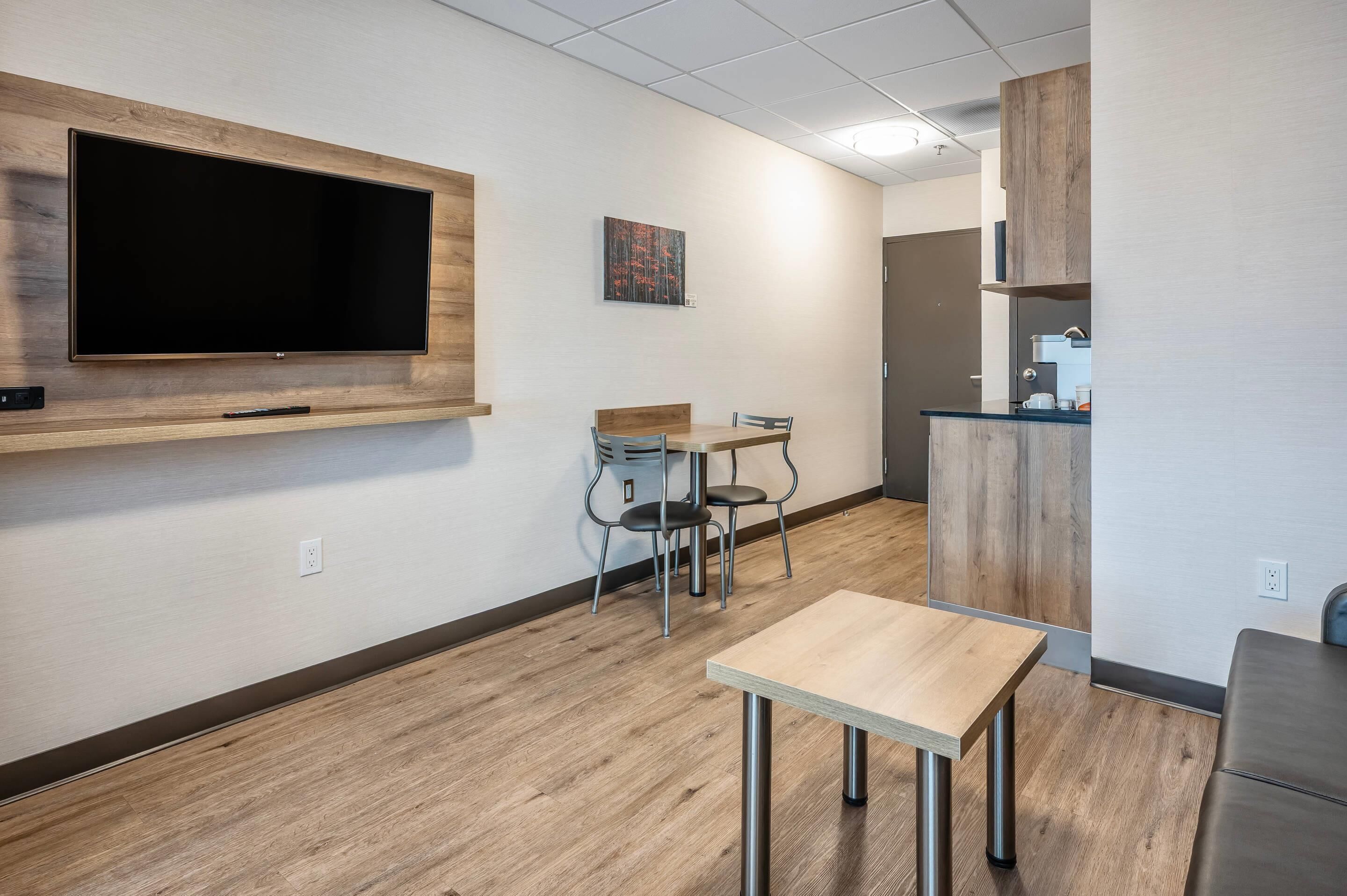 A modern office space with a large flat-screen TV mounted on a wooden panel, a small table with two chairs, and a kitchenette area with a coffee machine.