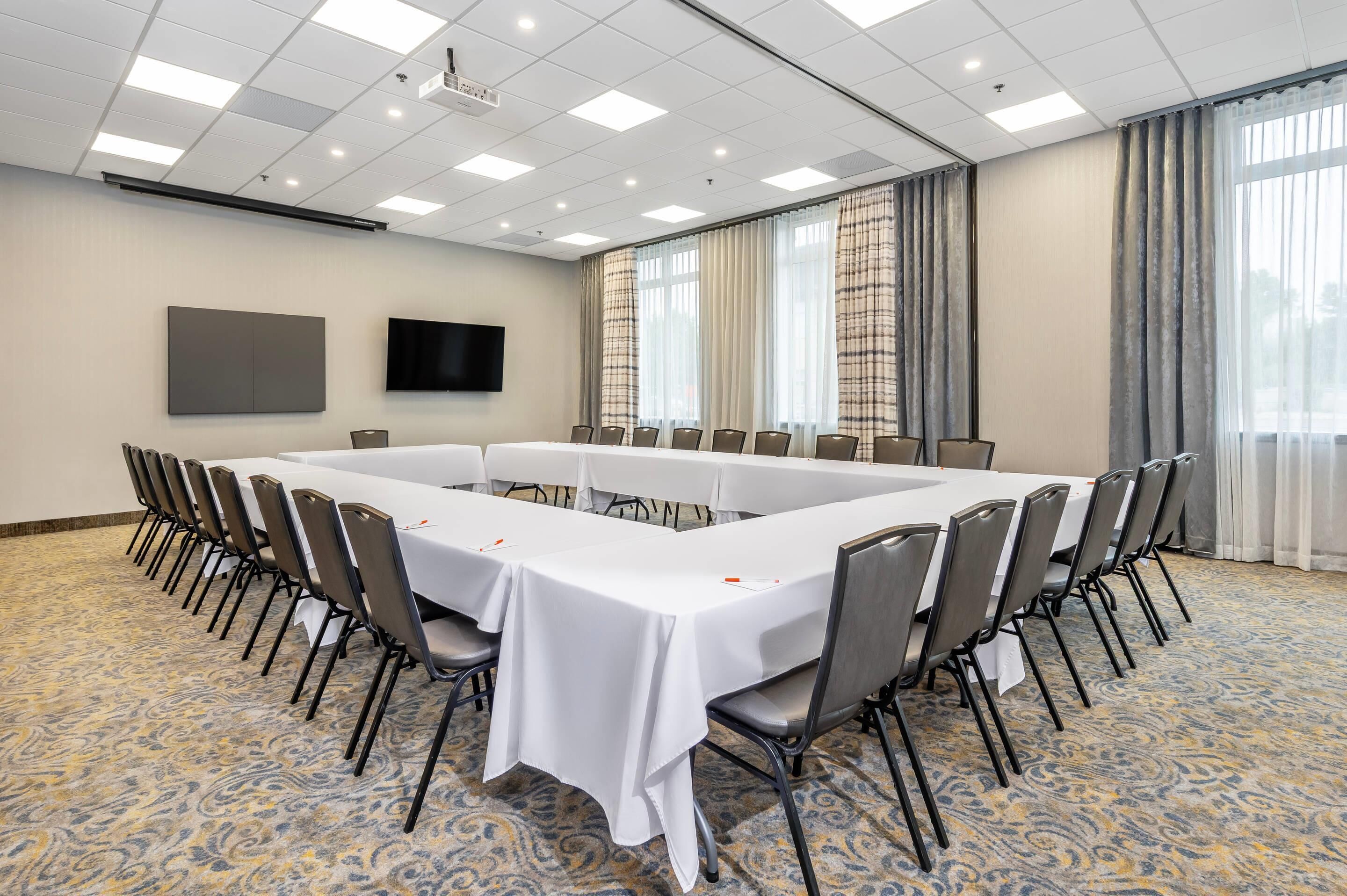 Un salón de conferencias con una mesa larga, sillas, un televisor de pantalla plana y ventanas grandes con cortinas.
