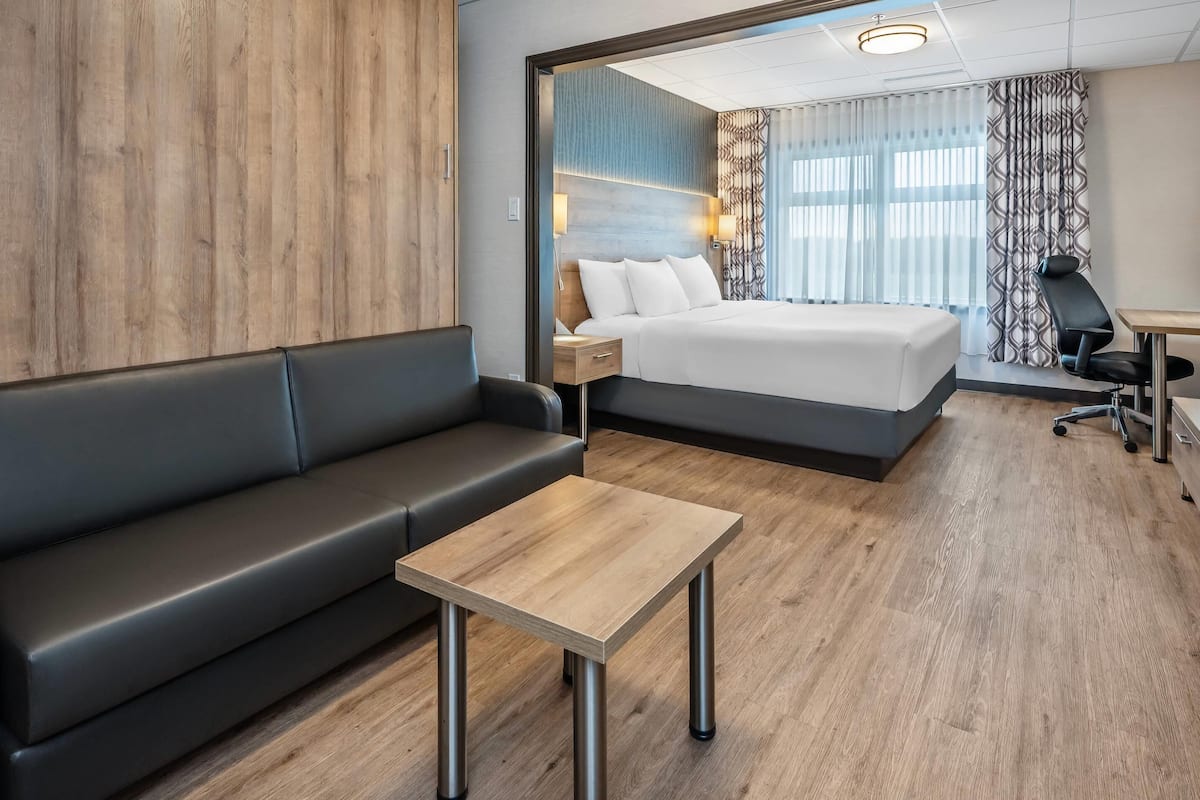 Una camera d'albergo moderna con pavimento in legno, un divano in pelle nera, un tavolo in legno, un letto con lenzuola bianche, una scrivania con una sedia e una tenda fantasia.