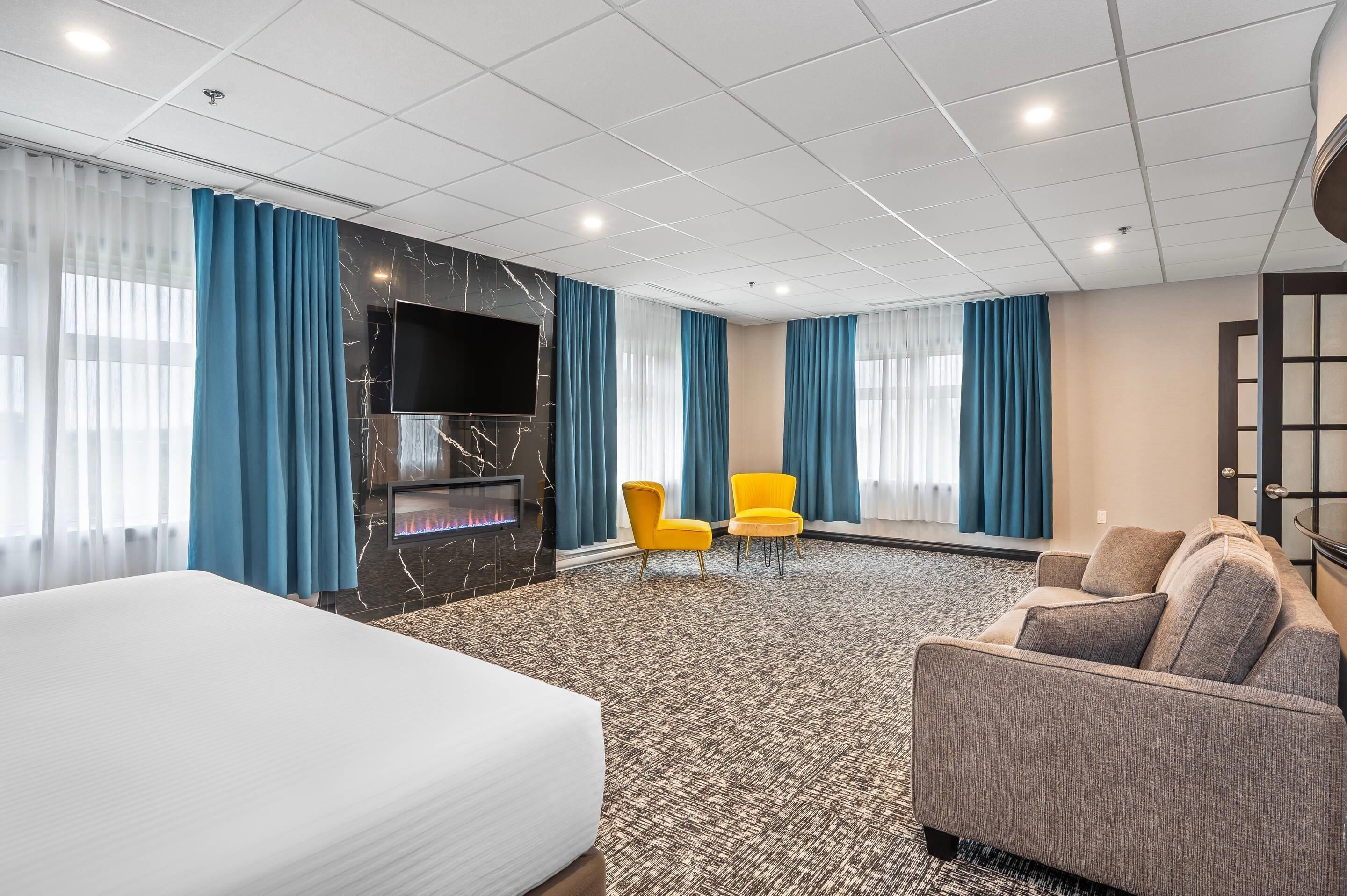 Habitación de hotel con una cama grande, un televisor de pantalla plana montado en una pared de mármol, dos sillas amarillas, un sofá gris y una alfombra estampada.