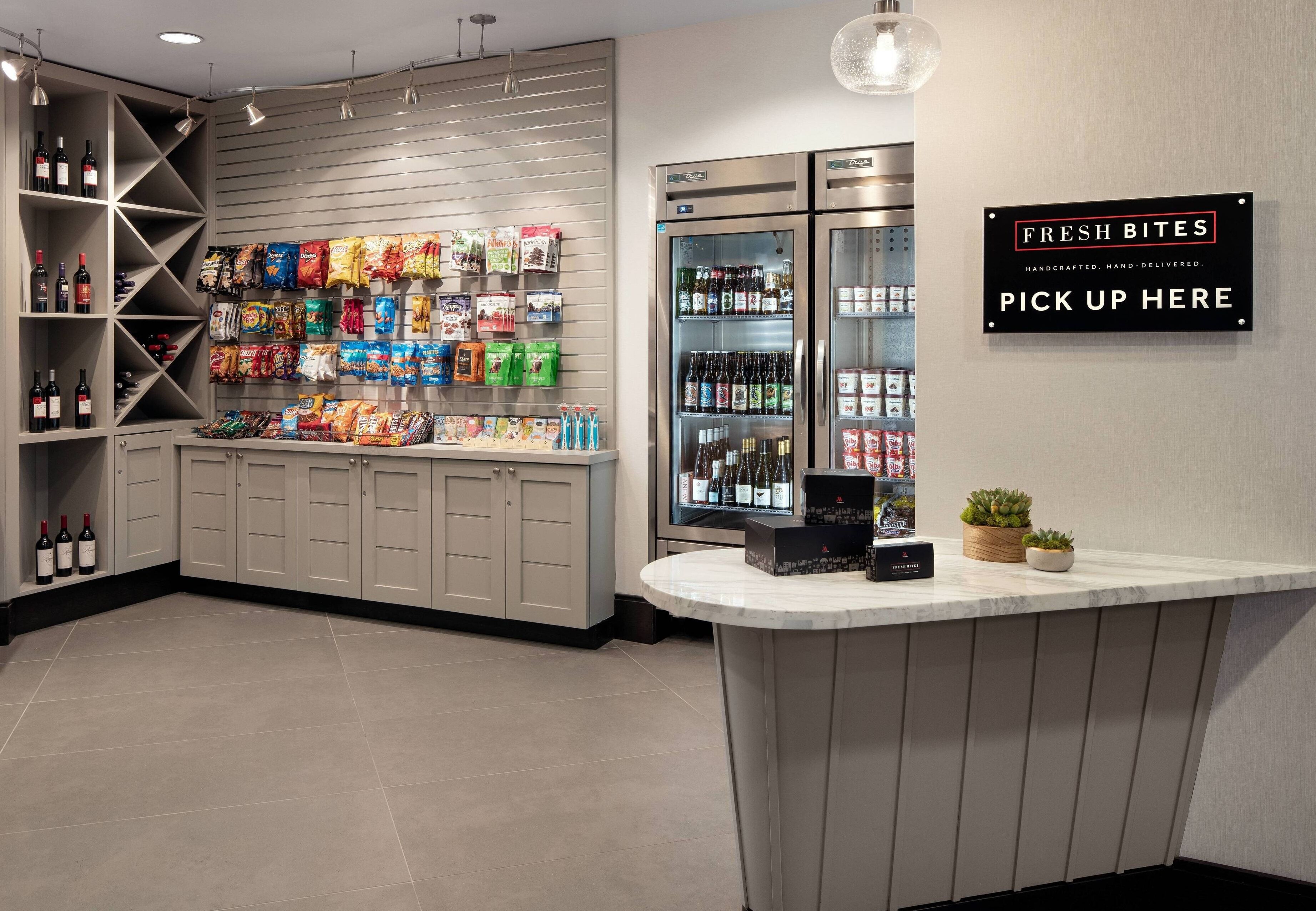 A well-lit snack bar with shelves stocked with snacks and beverages, a refrigerated display case, and a sign indicating a pick-up area for fresh bites.