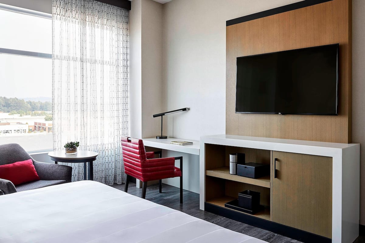 A modern hotel room with a large bed, a desk with a flat-screen TV, a red chair, and a view of the city.
