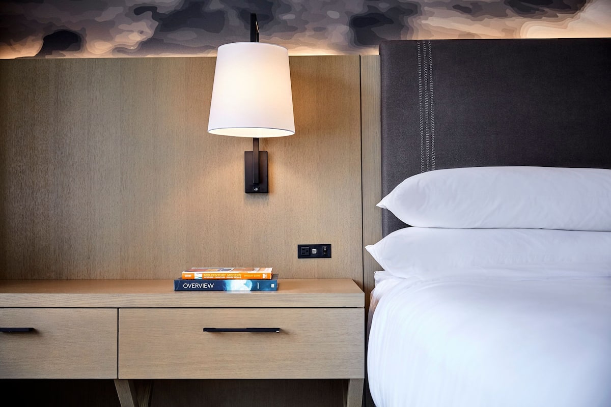 A neatly made bed with white linens, a bedside lamp, and a nightstand with a book titled "OVERVIEW".