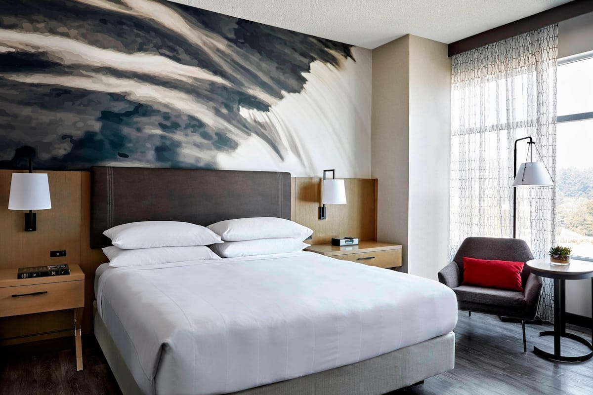 A modern hotel room with a large bed, two bedside tables with lamps, a small armchair with a red cushion, and a view of the outdoors through a patterned curtain.