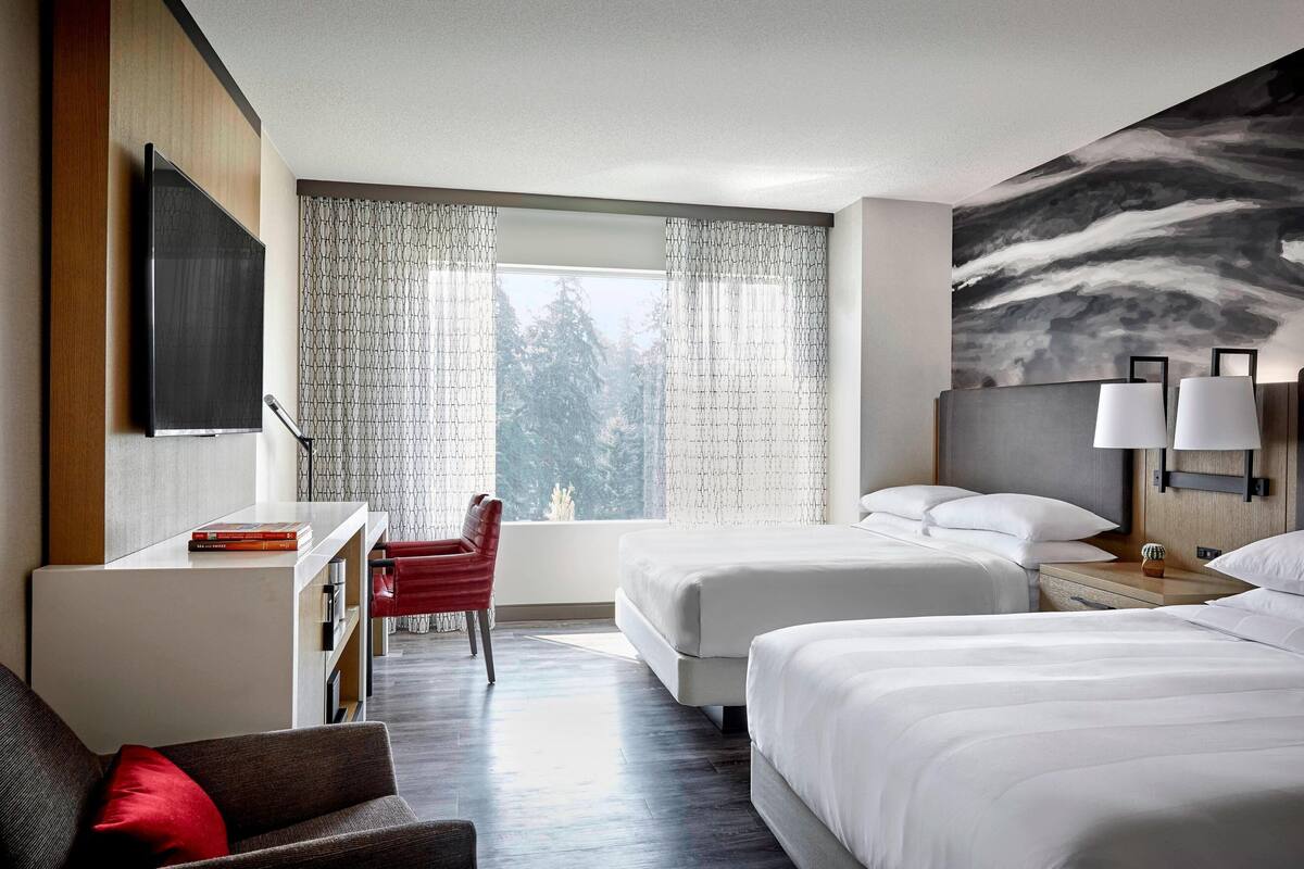 A hotel room with two beds, a desk, a chair, a TV, and a large window with curtains.