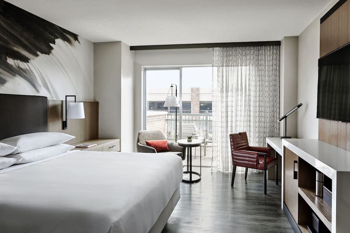 A modern hotel room with a large bed, a desk with a chair, a small table with a lamp, and a view of the city through large windows.
