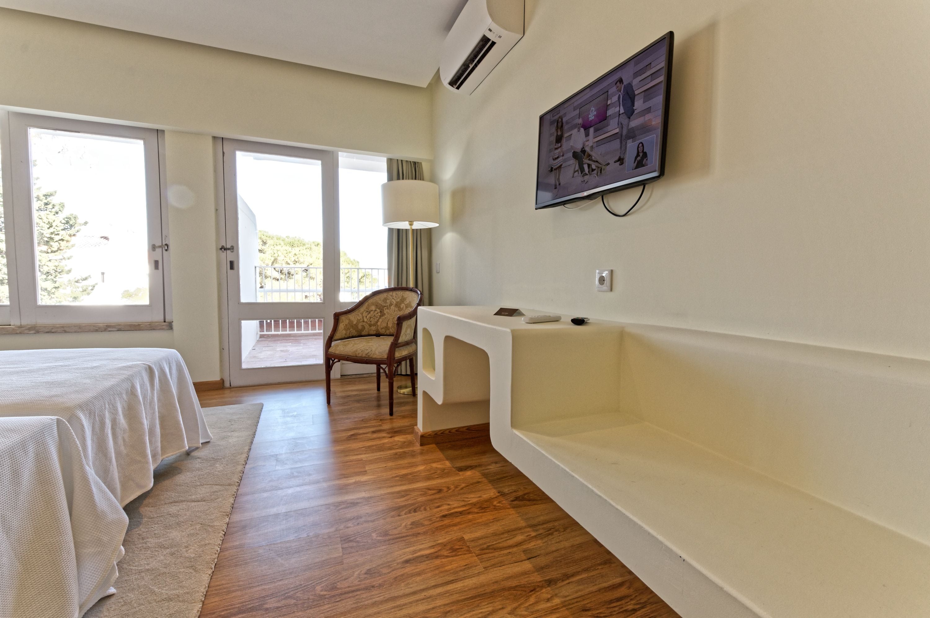 Un dormitorio con una cama, un escritorio, una silla, un ventanal grande y un televisor montado en la pared.