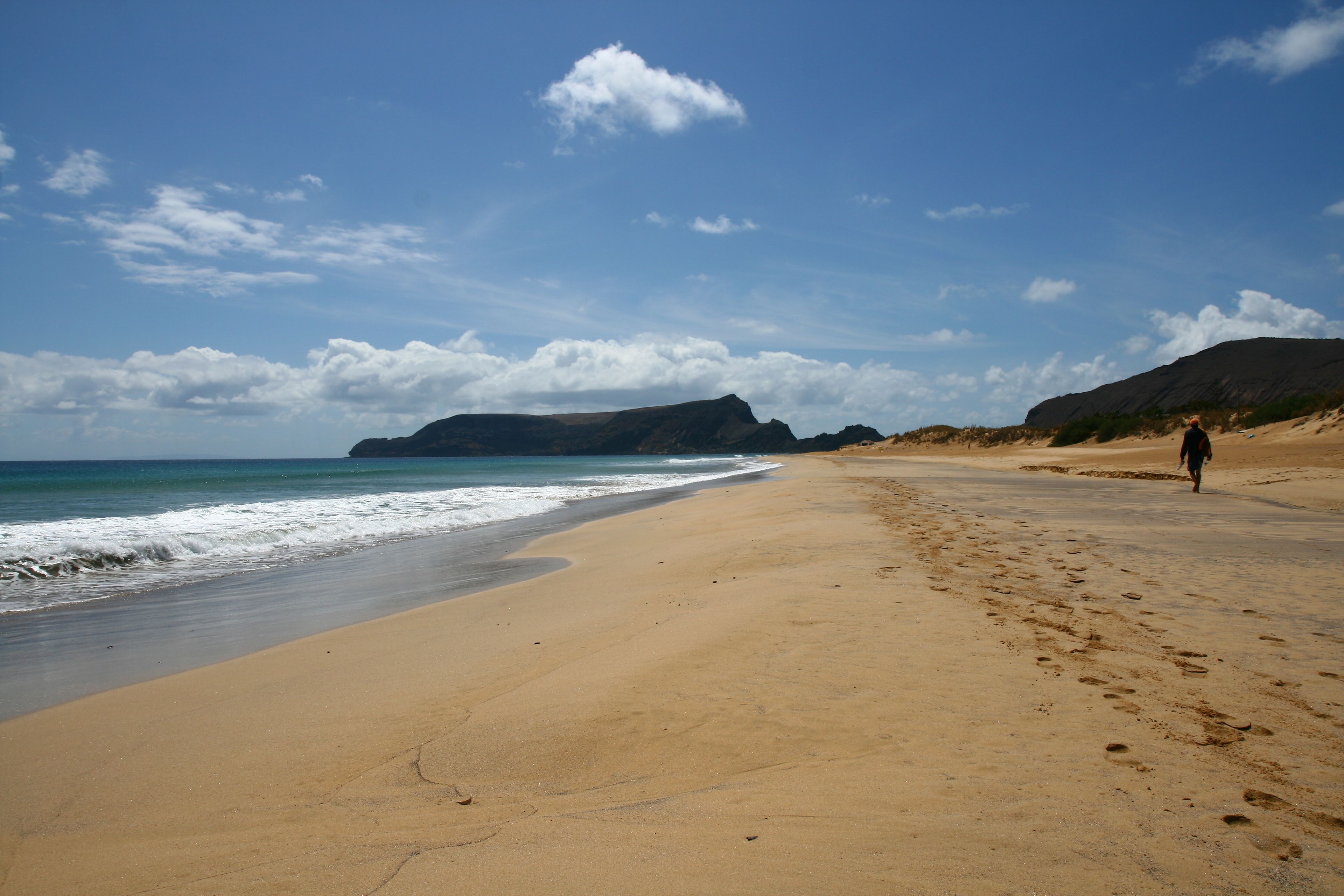 Una playa con una persona caminando, cielos despejados y montañas a lo lejos.