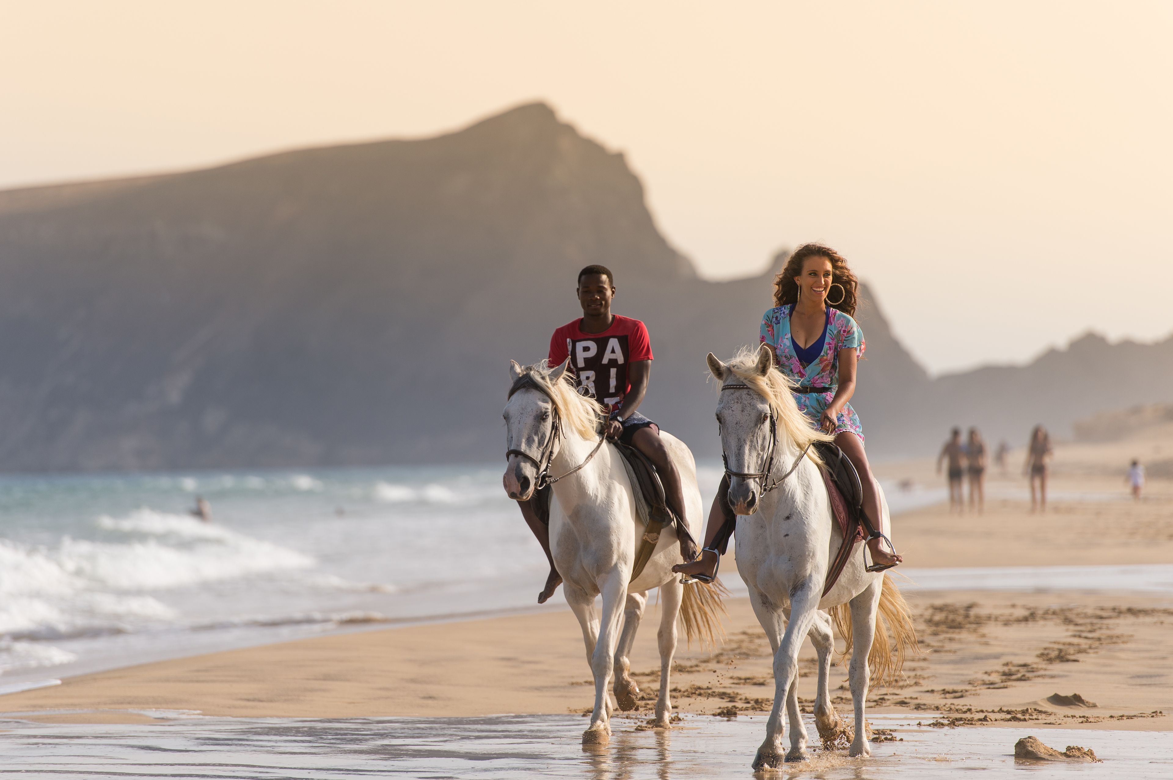 Dos personas montando a caballo en una playa, con una montaña al fondo.