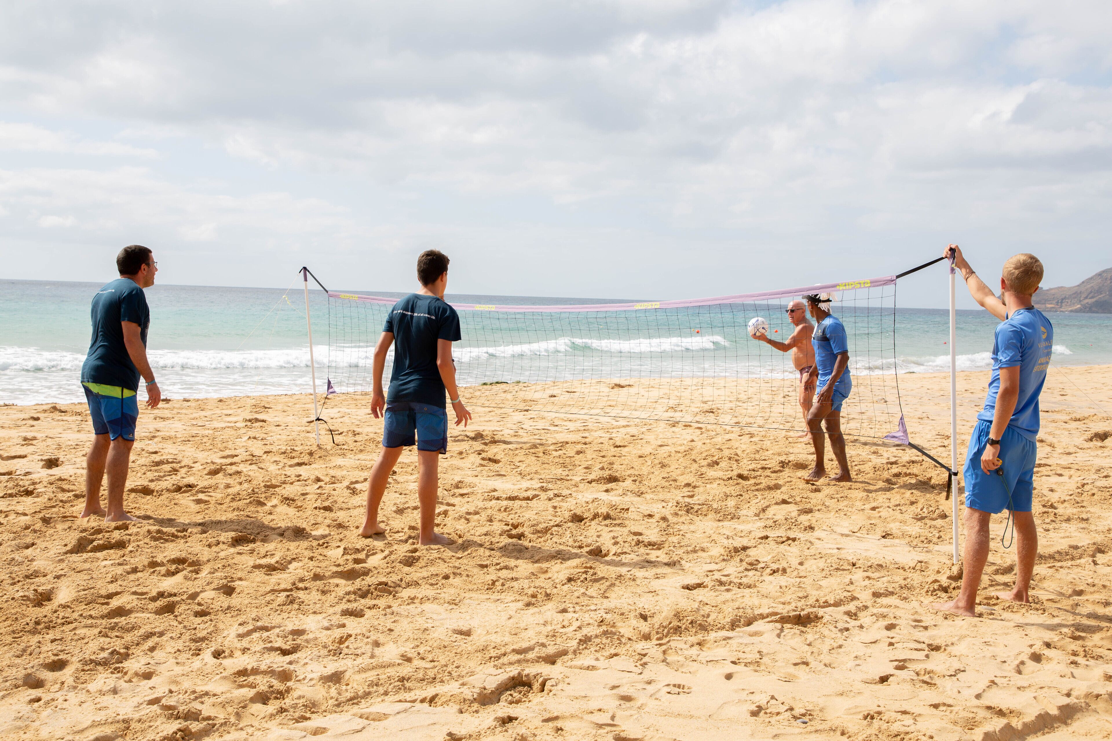 Partido de voleibol de playa con jugadores vestidos de azul y uno con una camisa oscura.