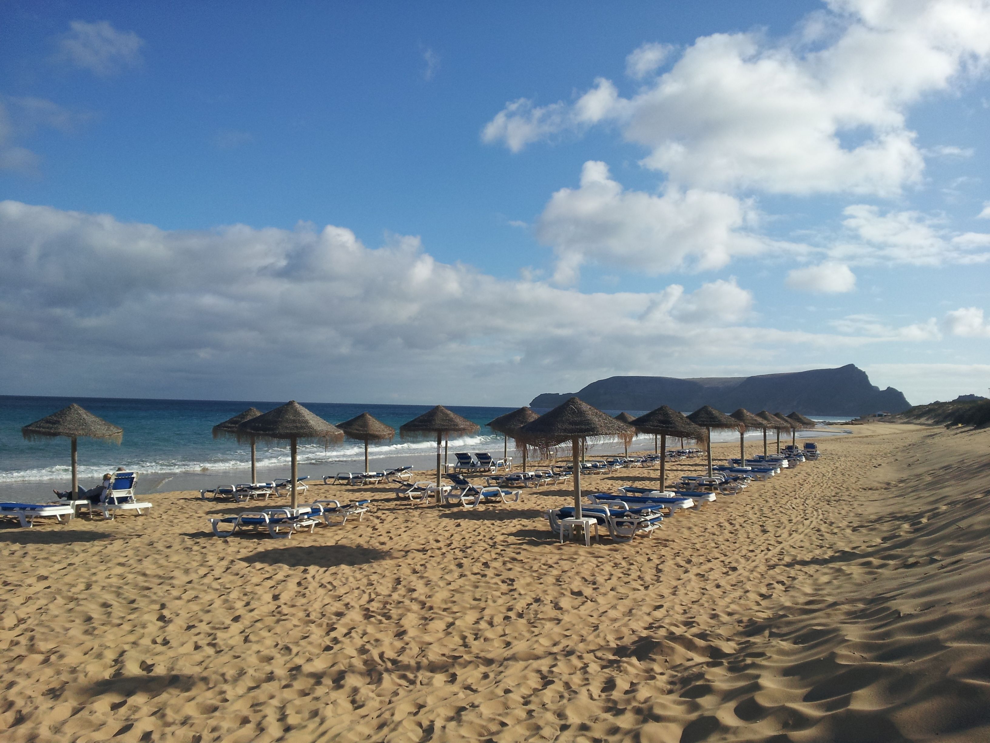 A beach with rows of sun loungers and thatched umbrellas, a clear sky, and a mountain in the background.