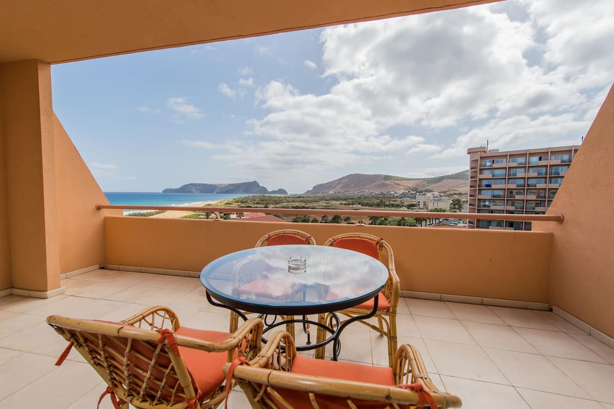 A balcony with a glass table and wicker chairs, overlooking a beach and mountains.