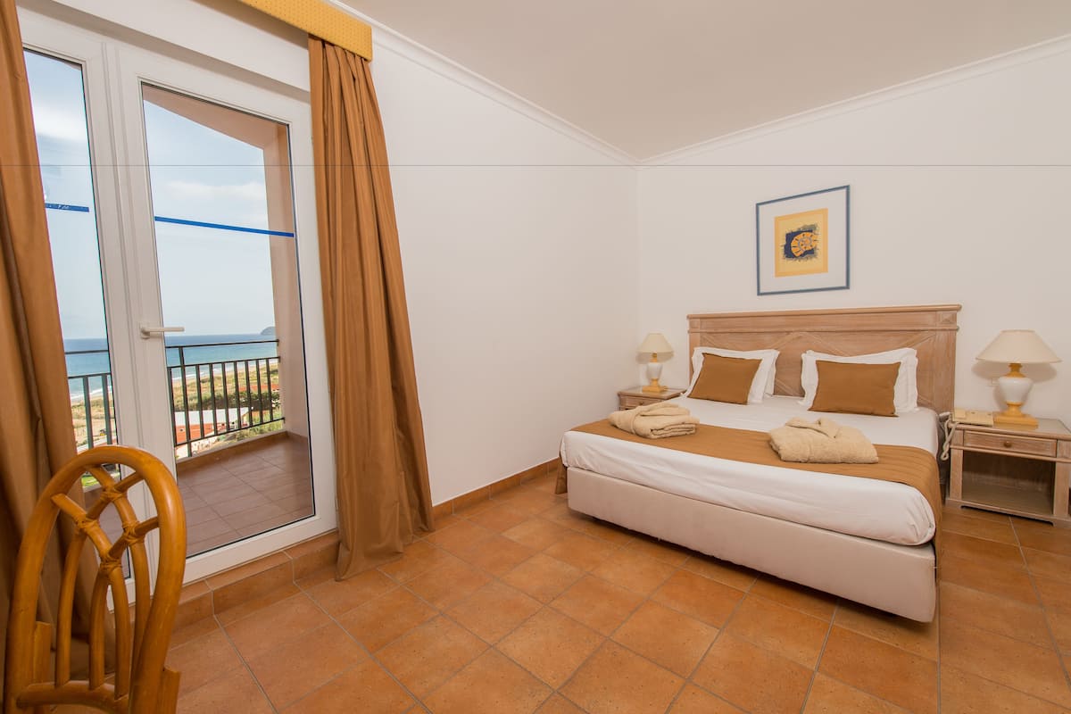A hotel room with a bed, a nightstand, a chair, a window with curtains, and a view of the sea.