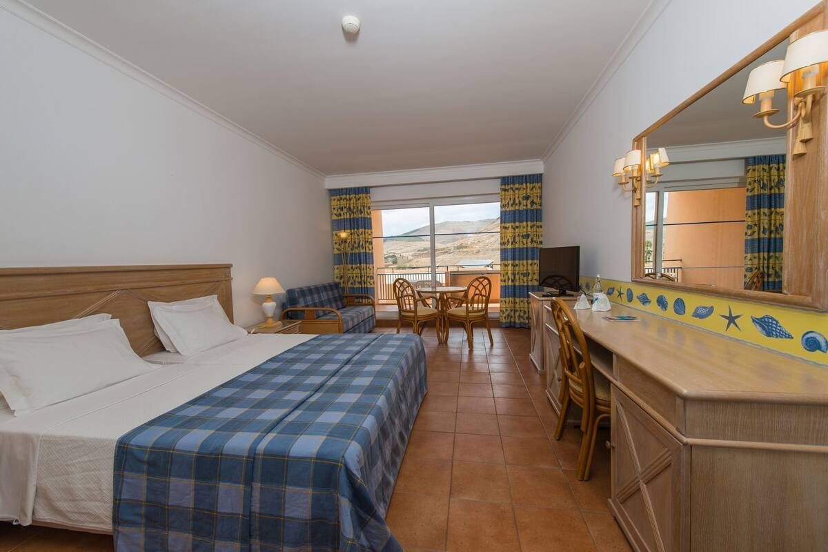 A hotel room with a bed, a desk, a chair, a TV, and a view of the landscape.