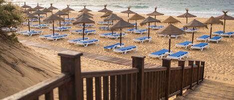 A beach with rows of sun loungers and thatched umbrellas, a wooden boardwalk, and the ocean in the background.