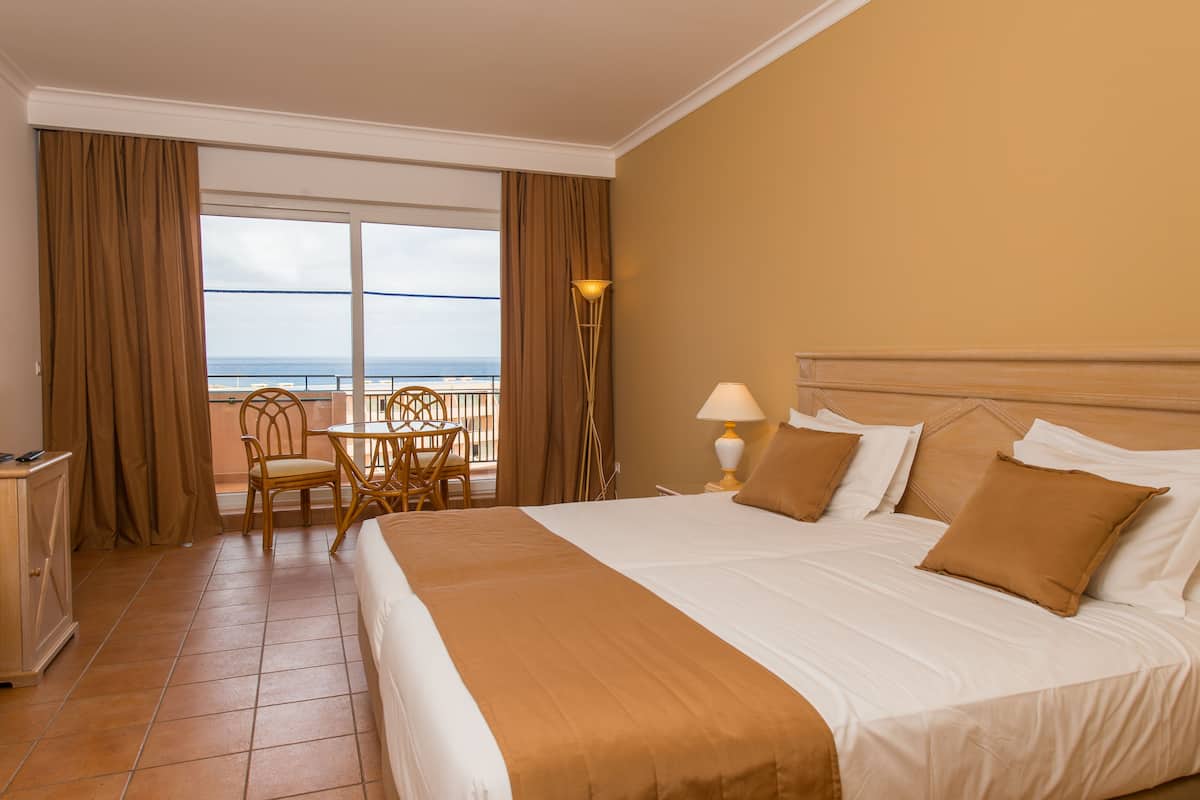 A hotel room with a large bed, a balcony with a view of the sea, a small table with chairs, and a lamp.