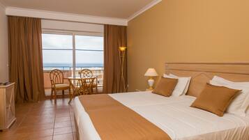 Camera d'albergo con un letto grande, un balcone vista mare, un tavolino con sedie e una lampada.