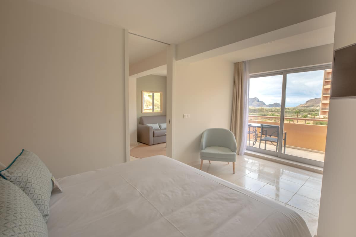 A hotel room with a large bed, a chair, a balcony with a view of the desert and mountains.